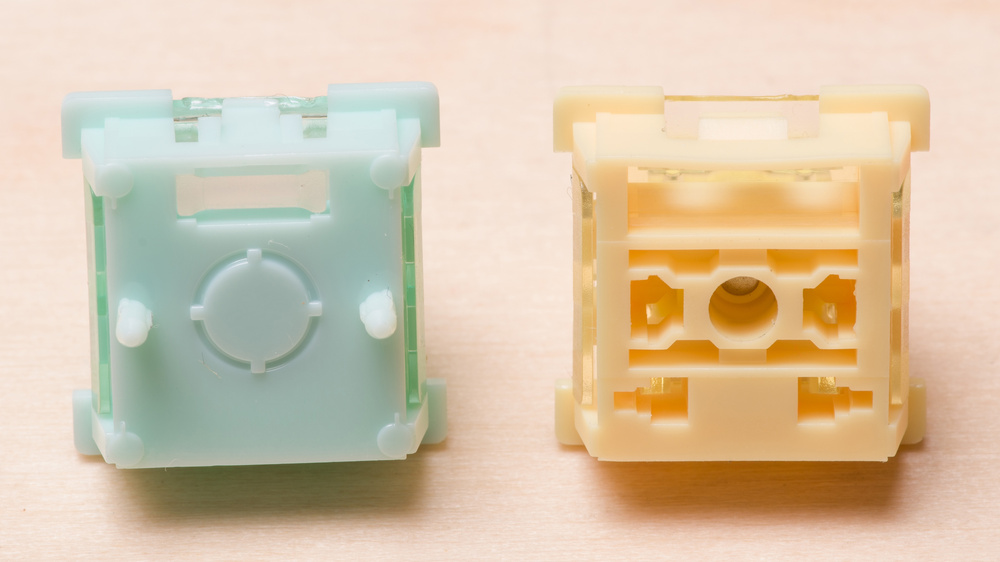 Two Hall effect keyboard switches housings displayed side by side on a light wooden surface. The left switch features a translucent mint green with a circular actuator stem in the center and two side stabilizing pins. The right switch has a honey-colored housing revealing internal orange components and mechanisms.