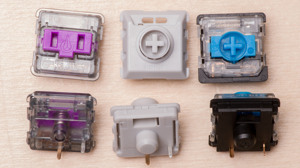 Six low-profile mechanical keyboard switches displayed in two rows of three on a light wooden surface. Top row shows a purple Kailh switch with clear housing, a white switch with a plus symbol, and a blue switch with clear housing. Bottom row displays a purple switch with clear housing and visible spring mechanism, a white switch with exposed actuator stem, and a black switch with clear housing showing internal components. All switches have their metal differently placed contact pins visible at the bottom.