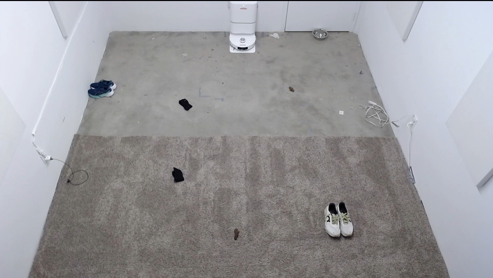 A photo of a room, with carpet covering half, with a robot vacuum in its dock at the center of the far wall. Shoes, socks, a USB cord, an extension cord, a pet bowl, and two deposits of pet waste are all strategically placed in the room.