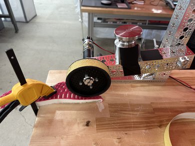 Abrasion wheel wrapped in sandpaper grinding a cut running shoe outsole held down by a bar clamp, mounted on a weighted aluminum frame for durability testing.