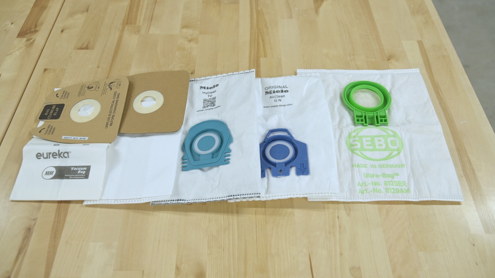 5 vacuum bags on a table. From left to right, the Eureka Mighty Mite bag, a Eureka Mighty Mite HEPA bag, a Miele TU bag, a Miele GN bag, and a Sebo D4 bag.