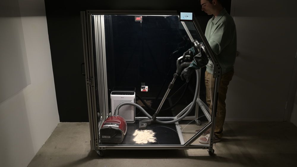 An acrylic box framed with extruded aluminum, measuring 1.5m tall, 75cm deep, and 1.2m long. There is a vacuum cleaner inside with dust on the ground. The vacuum operator uses through-gloves to manipulate the vacuum wand.
