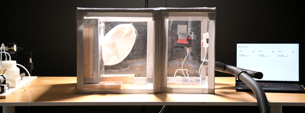 An acrylic box on a table with a nozzle on the left and a vacuum cleaner tube on the right. There is a vacuum bag inside the box.